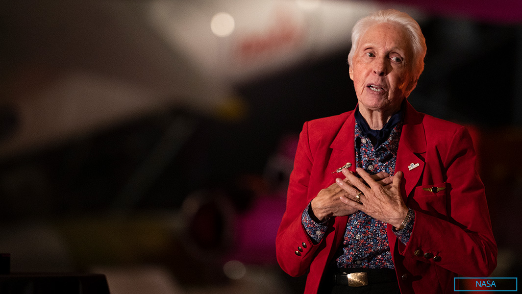 wally funk wearing a read blazer and giving a speech